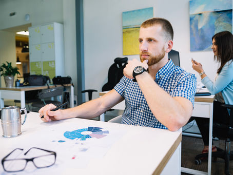 Man in blue checkered shirt thinking at desk with charts; woman using phone in background. Office setting, colorful art on walls.