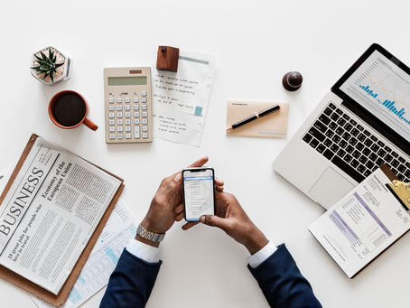 Man in suit holding phone at a desk with newspaper, laptop, chart, calculator, notepad, pen, coffee, and plant, creating a busy office vibe.