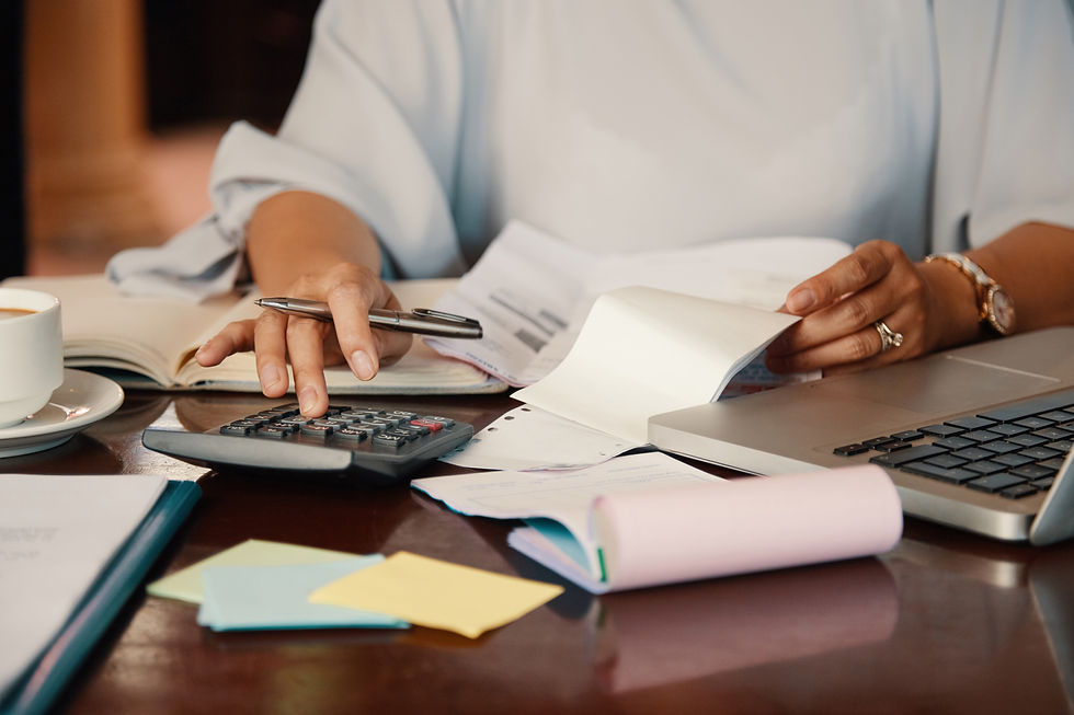 Hands using a calculator and flipping through a notebook on a cluttered desk with a laptop, papers, sticky notes, and a coffee cup.