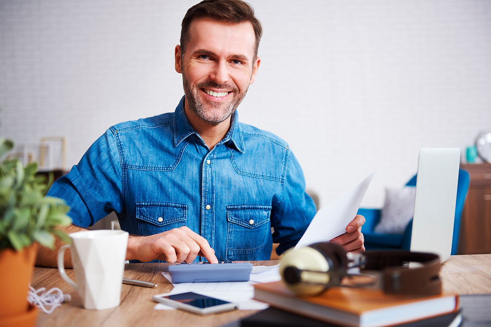 Smiling man in denim shirt works at desk with calculator and papers. Laptop, coffee mug, and headphones present in cozy office setting.