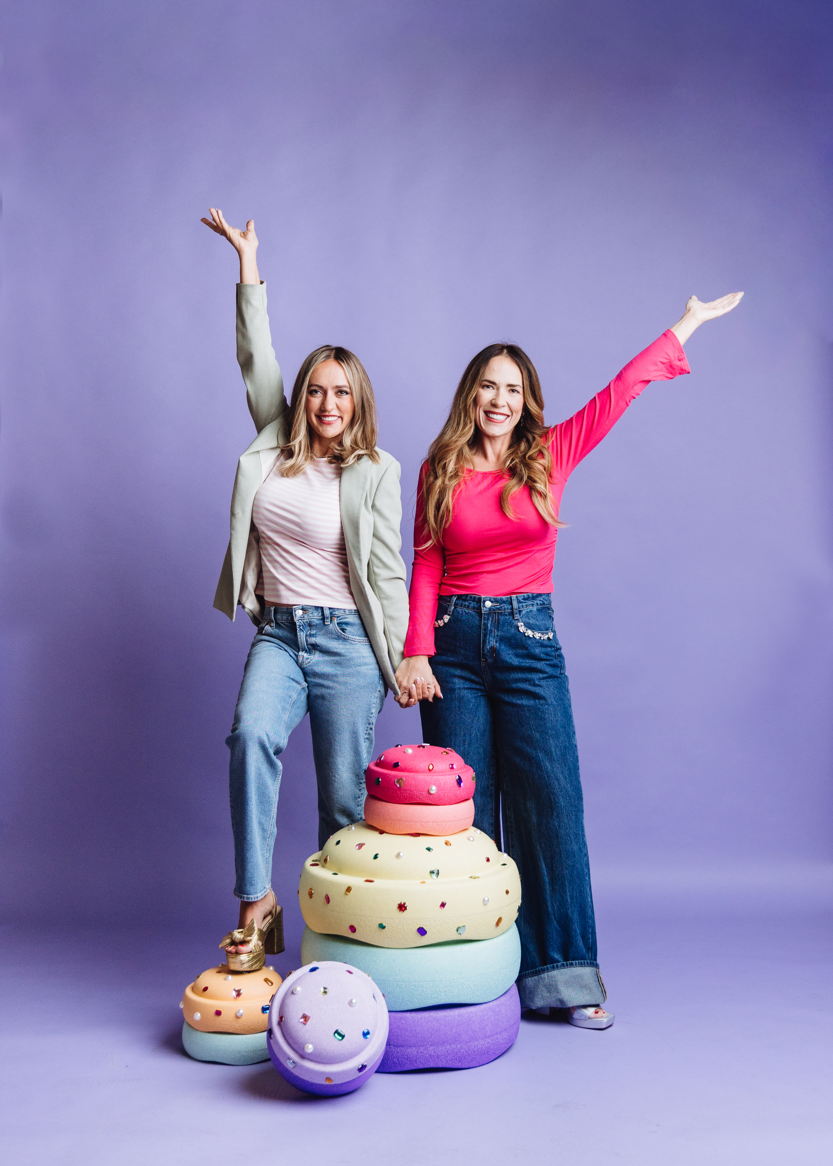 Mandy and Mel celebrating with their arms up next to a stack of toy cakes
