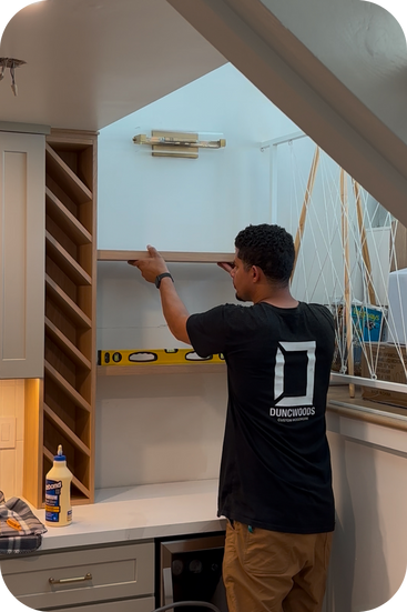 Christopher Duncan wearing a black t-shirt with a Duncwoods logo. He is installing a floating shelf and wine bar.  There is a level, wood glue and miscellaneous tools present.