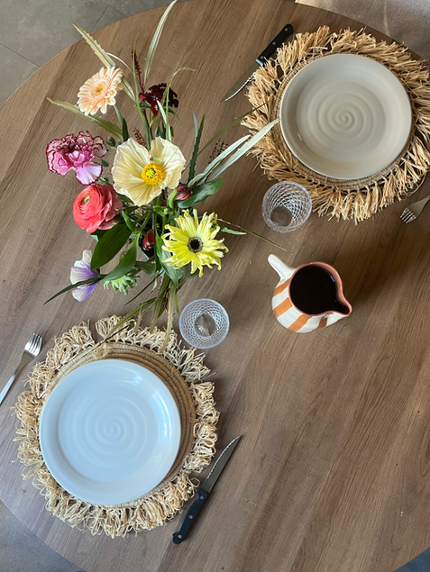 Table vue du dessus avec deux assiettes beige un carafe rayée marron et blanc et des sets de table en paille et un bouquet de fleur multicolor le tout chalereux