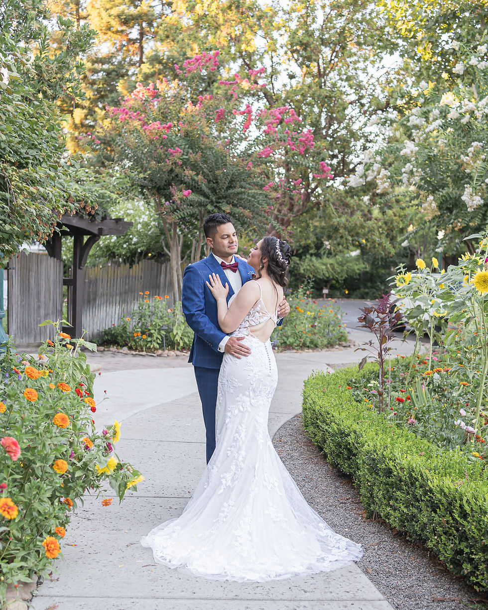 A bride in a white lace gown and a groom in a blue suit share a romantic moment in a flower-lined garden at Wine and Roses in Lodi. The couple stands on a sunlit path surrounded by vibrant flowers and greenery, captured during their outdoor wedding ceremony.