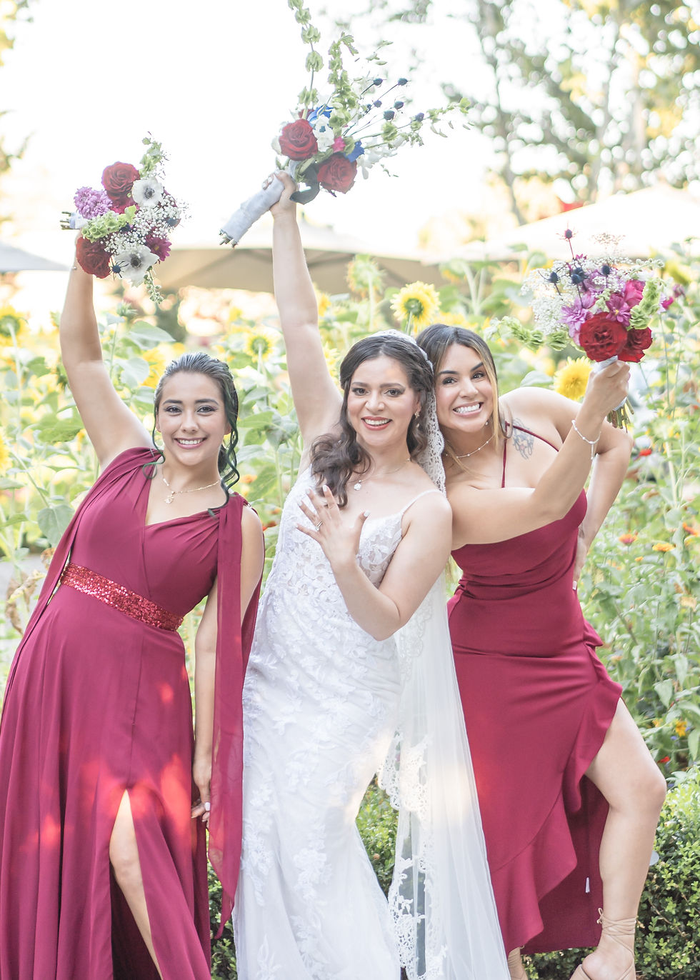 Three women in formal attire, including a bride in a white lace dress and bridesmaids in red dresses, celebrating in a garden with sunflowers at Wine and Roses in Lodi, CA.