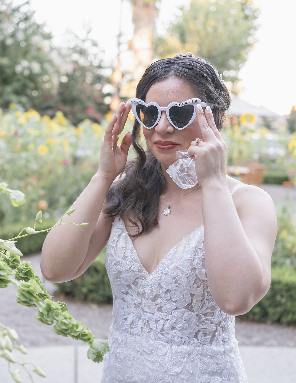 Bride in a white lace dress adjusting sunglasses in a lush garden at Wine and Roses in Lodi, CA, with a pendant necklace and vibrant flowers in the background.