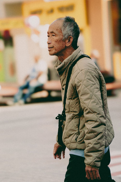 Elderly Asian man walking across a street, wearing a puffy jacket.