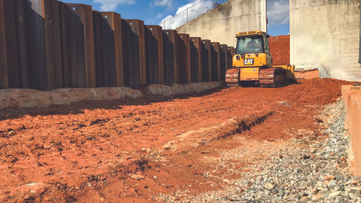 Steel sheet pile wall alongside compacted red earth and a dozer, used for earth retention during construction near large concrete abutments.