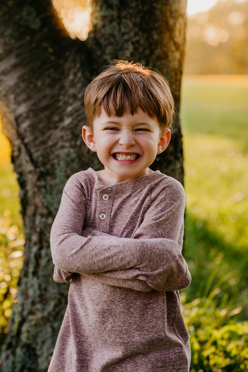 Four year old boy with brown hair stands in front of a tree truck at sunset and smiles big at the camera in Cincinnati, Ohio.