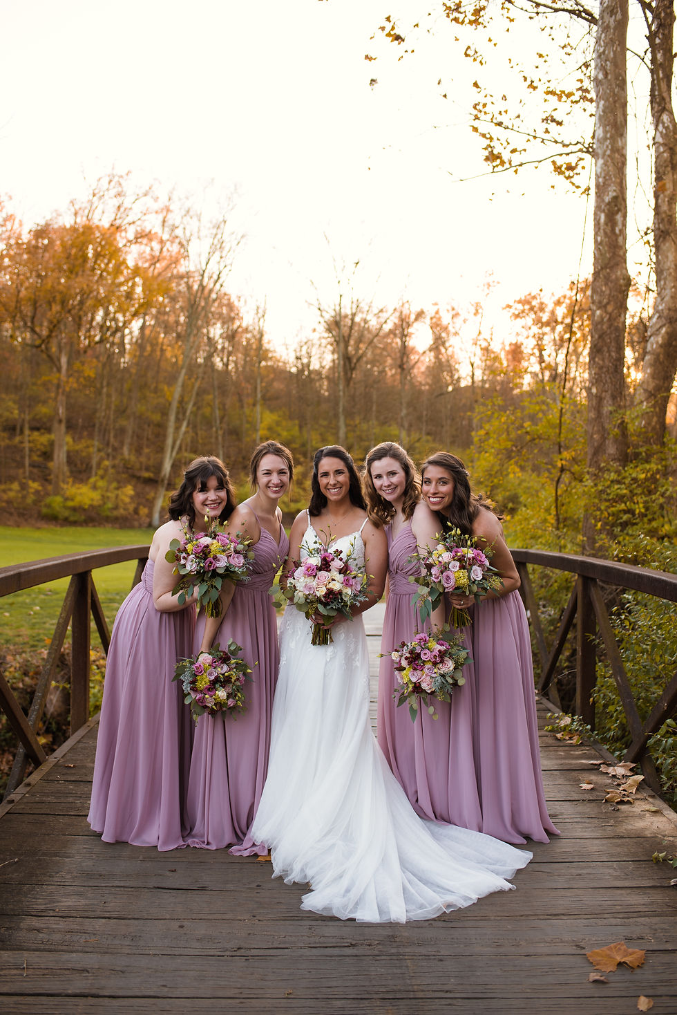 Bride in white dress is surrounded by four bridesmaids in blush pink dresses as they huddle together and smile at the camera on a bridge at sunset in Cincinnati, Ohio.