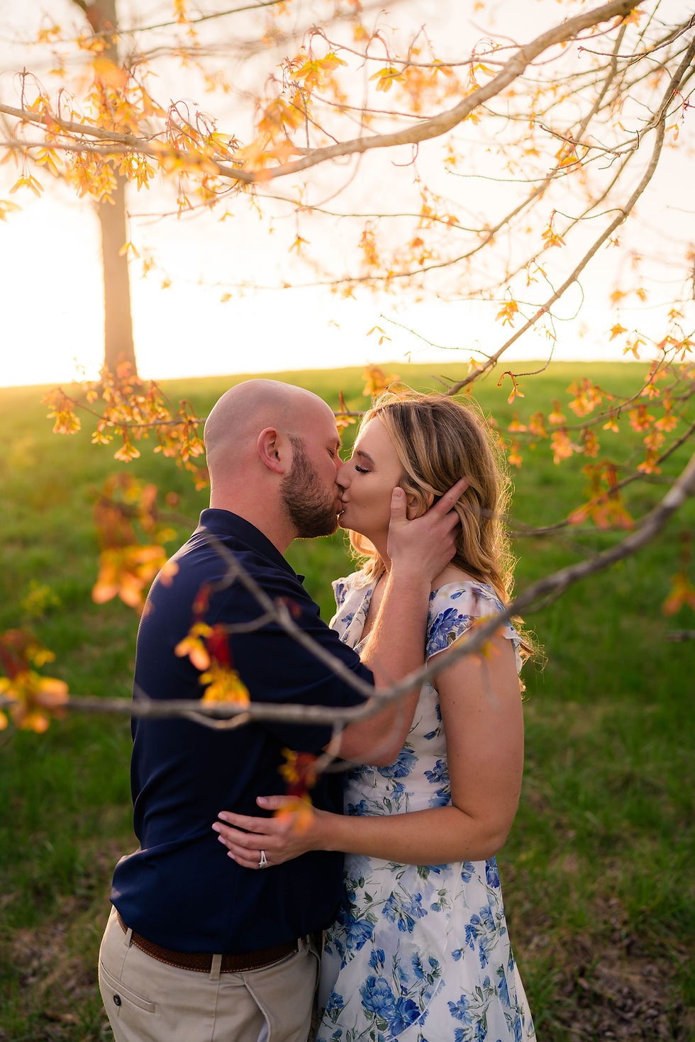 A couple kisses under a tree with yellow leaves. The woman wears a floral dress. The setting sun creates a warm glow on a grassy field.