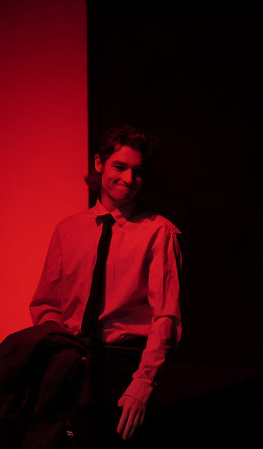 A man in a dress shirt stands on a stage in red light. There is a white door behind him that looks red in the light.