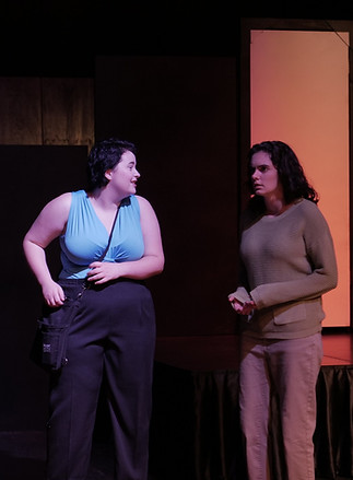 Three actors on stage. One wears all black behind a bar. The other two talk to each other on the right.