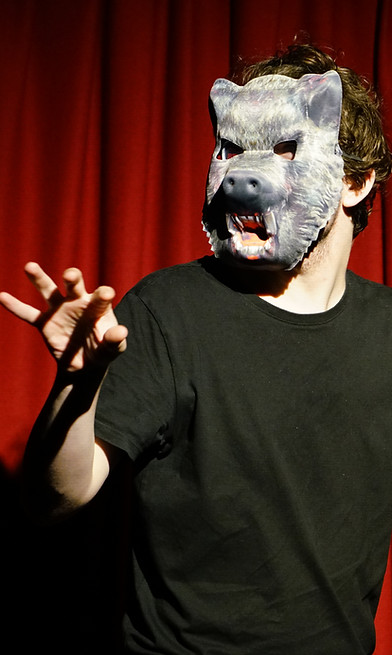 A man stands on stage. He is wearing all black with a grey werewolf mask.