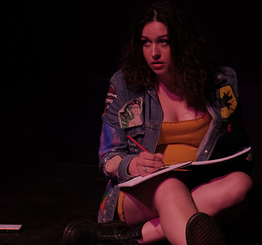 An actor sits on the ground drawing. She has a yellow dress on and a denim jacket.