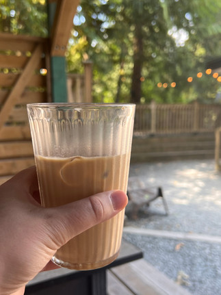 A white-skinned hand holding up a ribbed glass of milky coffee in front of the forest backdrop