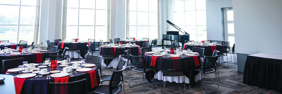 Photograph of the Fitton Center's Vista Room. The room holds many tables with layered black and white linens and red napkins. The room is brightly lit by a row of floor-to-ceiling windows. In the back corner a piano can be seen propped open.
