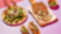 Sliced roast pork on wooden board beside a green salad, baguettes, and a herb sauce on a pink and lavender table.