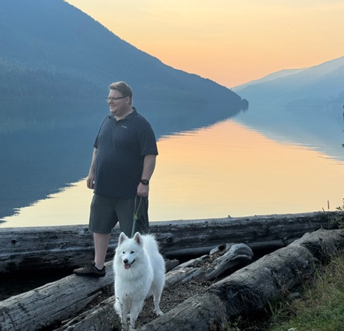 Man and white dog by a calm lake with mountains at sunset.