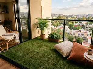 Terraza acogedora con césped artificial, plantas y cojines; vista panorámica de la ciudad al atardecer.