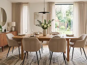 Comedor moderno y luminoso con mesa servida, sillas beige, flores blancas y vista al jardín tras cortinas claras.