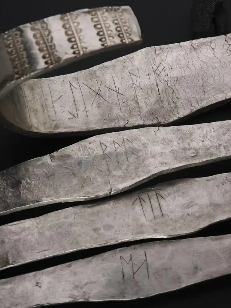 Livros Vikings | Close-up de pulseiras/placas metálicas gravadas com símbolos rúnicos, sobre fundo preto, em tom cinza envelhecido.