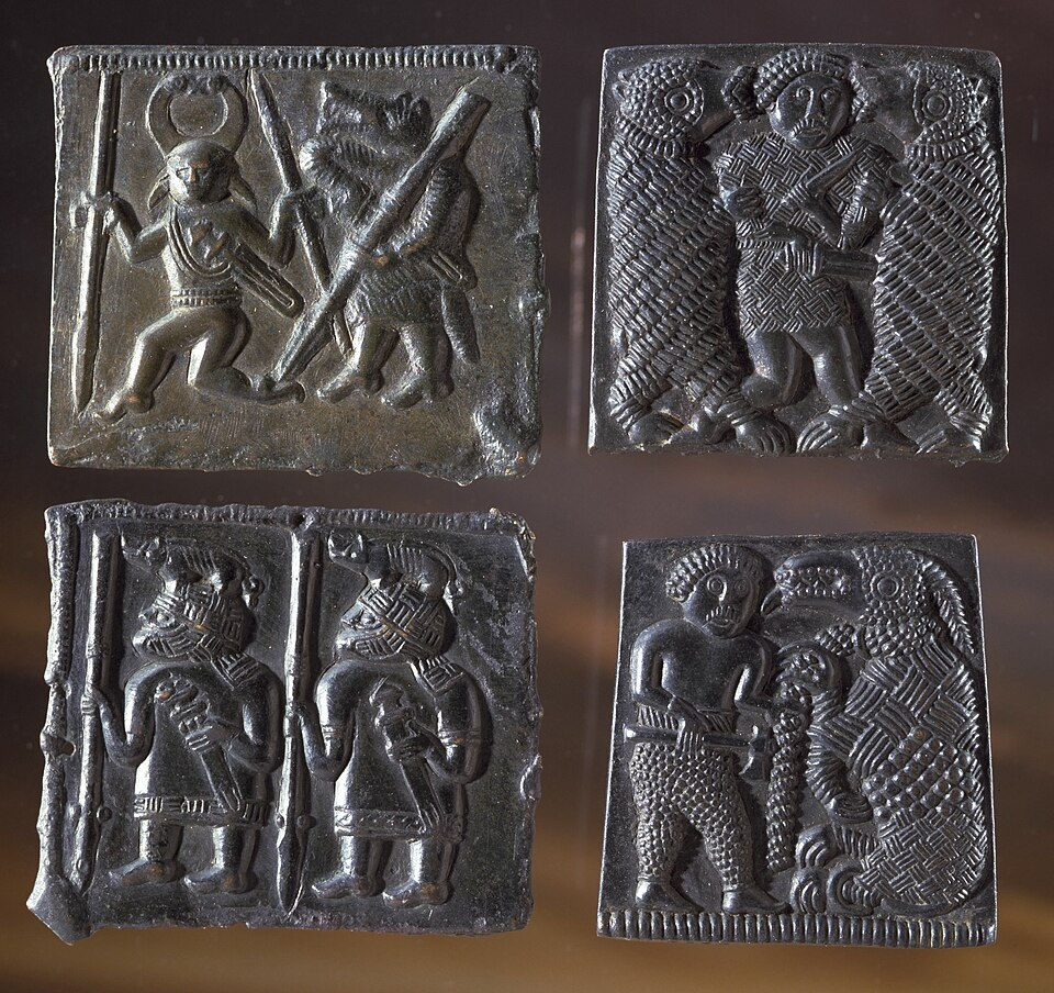 Livros Vikings | Quatro placas metálicas em relevo com guerreiros e figuras animais escuras, em fundo desfocado, com aspecto antigo.