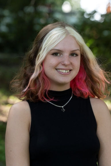 Smiling person with two-tone hair stands outdoors. Wears a black top and necklace. Background is a blurred, leafy setting.