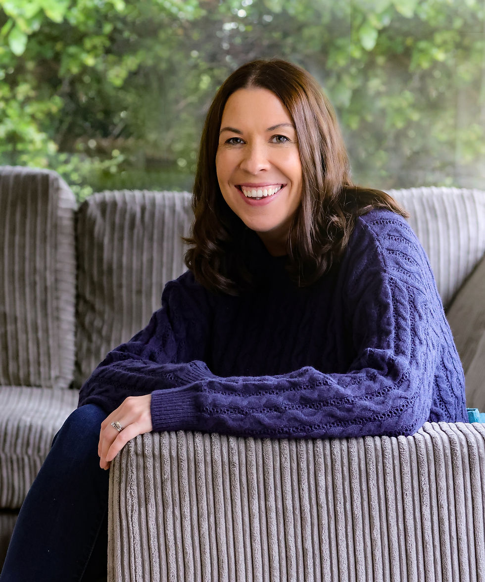 A smiling woman with shoulder-length dark brown hair is seated on a comfortable, striped, gray armchair. She is wearing a cozy navy blue sweater and has her left hand resting on the armrest, displaying a silver wedding ring. The background features large windows with a lush, green garden outside, creating a peaceful, natural atmosphere. Her relaxed posture and warm expression convey a sense of approachability and calm.