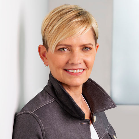 Seattle headshot of a smiling blonde woman with short hair in a gray jacket with a popped collar, leaning against a bright office wall.