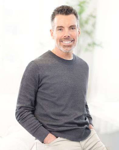 Relaxed three-quarter headshot of a smiling man with gray-flecked hair and beard, hands in pockets, wearing a charcoal sweater in a bright airy room.