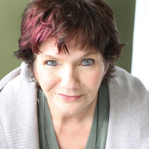Creative headshot of a smiling woman with short dark hair and pink highlights, wearing a gray cardigan against an olive-green background.