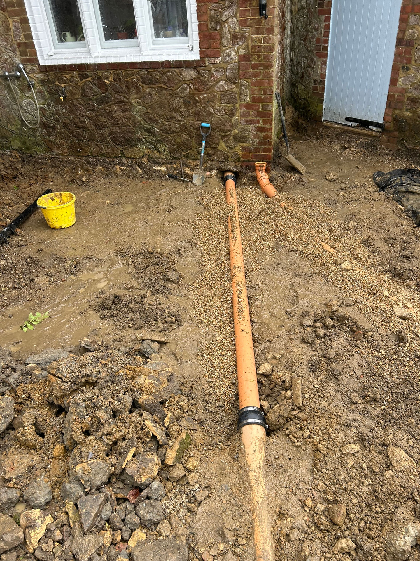 a yellow bucket sits in the mud next to a drain pipe
