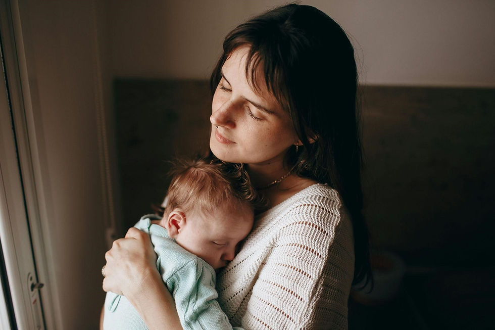 Woman in a white sweater holds a sleeping baby in a light blue outfit. She appears peaceful, with a soft, dimly-lit background.
