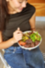Woman in black top and jeans smiling while holding a salad bowl with steak, tomatoes, and greens, seated on a woven chair indoors.