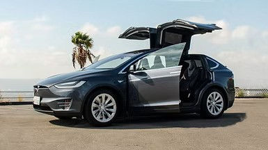 A matte black Tesla Model X parked with its signature falcon-wing doors and front doors open wide on a coastal road.