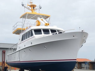 A large white boat with a yellow roof is on a dry dock, surrounded by a cloudy sky and industrial buildings. A person stands nearby.