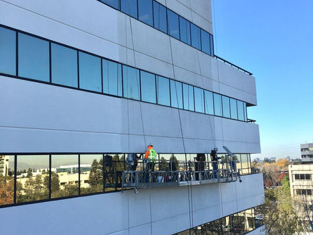 Window cleaners stand on a suspended platform cleaning a tall white building, with blue sky reflecting in the glass windows.