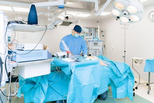 Surgeon in blue scrubs and mask operates in a well-lit operating room. Medical equipment surrounds the area. Calm, professional atmosphere.