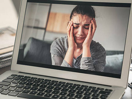Woman on laptop screen holding head in frustration. Background shows a cozy home setting with a blue couch. Mood is stressed.