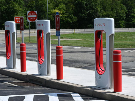 Tesla charging stations with red accents in a parking lot. Green trees in the background. "Do Not Enter" and other signs visible.
