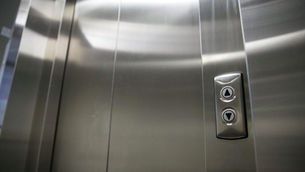 Close-up view of a clean, stainless steel elevator car door entrance with two metallic directional call buttons, showing well-maintained sill tracks free of summer debris.