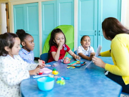 Children sit at a blue table with a teacher in a yellow sweater, engaged in a colorful craft activity. The setting has teal cabinets.