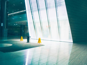 Bright, modern lobby with large glass panels, two yellow caution signs, and a trash bin. Glossy floors reflect blue and white hues.