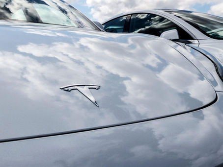 Close-up of a silver car hood with a Tesla logo, reflecting cloudy sky. A second car is partially visible. Bright, calm atmosphere.