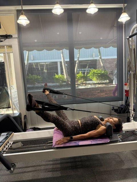 Woman doing a Pilates reformer exercise with straps in a bright studio, lying on a padded carriage near a large window.