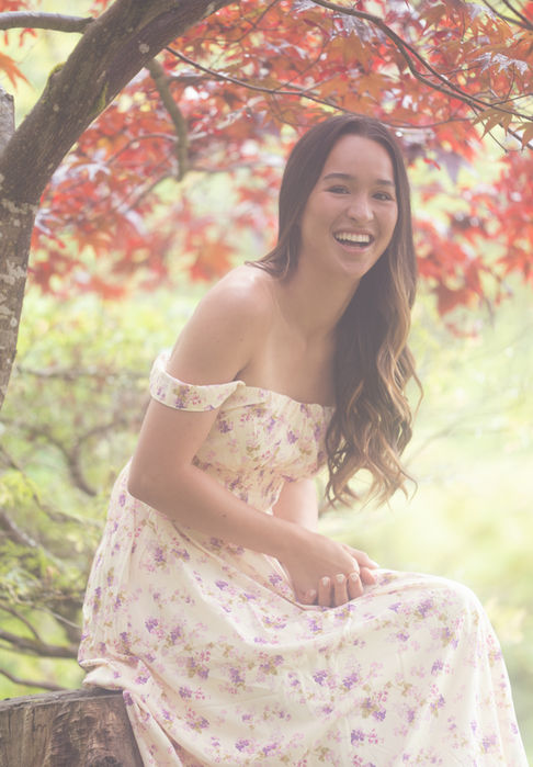 Woman in floral dress smiling under red-leaved tree.