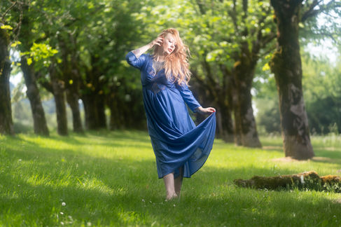 Woman in blue dress standing on grass, surrounded by trees.