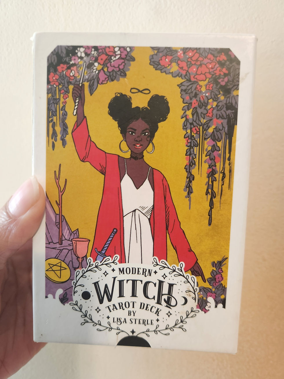 Box for Modern Witch Tarot by Lisa Steel shows a brown skinned woman holding a wand aloft. She is wearing a white dress and a reg cardigan and the page is decorated with many hanging flowers.