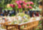 Outdoor wedding setup with wooden tables, pink flower centerpieces, and greenery. Guests in background under trees, creating a festive mood.